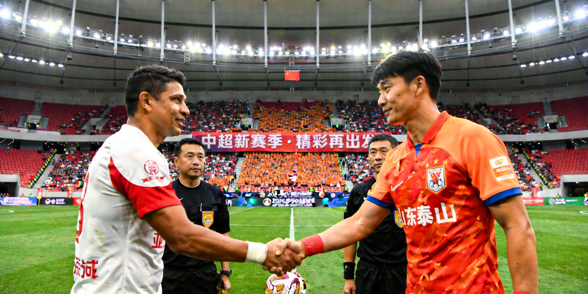 中超新赛季宣传横幅，上海海港与山东泰山球员在中圈开球前握手致意，背景是坐满观众的现代化足球场